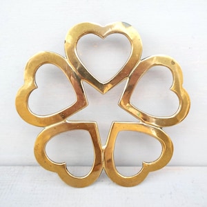 May include: A gold-colored trivet in the shape of a flower, with five heart-shaped petals arranged around a central star. The trivet is made of metal and has a shiny finish. The background is a white surface.