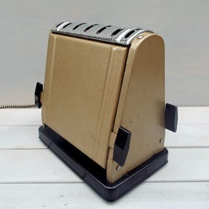 May include: A vintage gold-coloured toaster with black accents and a chrome top. The toaster has two levers on the side and a cord.
