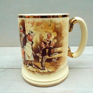 May include: Cream ceramic mug with gold detailing on the rim and handle. The mug has a sepia-toned illustration of a chef and children. The base of the mug reads "Oliver Asks for More".