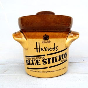May include: A ceramic Harrods Blue Stilton cheese crock with a two-tone brown and tan glaze. The front features the Harrods logo, a royal crest, and text reading "Finest Selected Blue Stilton Cheese" and "Harrods Limited Knightsbridge London England."