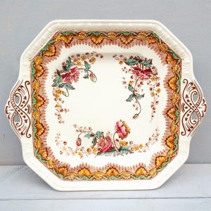 May include: An octagonal, off-white serving platter with a floral design. The rim features a decorative border of green, gold, and brown. The center of the platter displays pink and red flowers with green leaves. Handles are on each side.