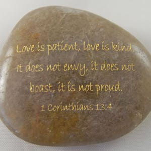 May include: A smooth, oval-shaped stone with a light brown hue. Gold lettering displays the quote "Love is patient, love is kind..." from 1 Corinthians 13:4. The stone's surface has subtle variations in color and texture.