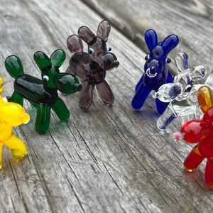 K&ouml;nnte beinhalten: Eine Sammlung farbenfroher Glas-Hundefiguren in Gelb, Gr&uuml;n, Lila, Blau, Klar und Rot. Die Miniatur-Skulpturen sind auf einer verwitterten Holzoberfl&auml;che angeordnet und zeigen ihre leuchtenden Farben und ihr verspieltes Design. Jeder Hund hat eine einzigartige Farbe.