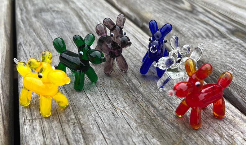 K&ouml;nnte beinhalten: Eine Sammlung farbenfroher Glas-Hundefiguren in Gelb, Gr&uuml;n, Lila, Blau, Klar und Rot. Die Miniatur-Skulpturen sind auf einer verwitterten Holzoberfl&auml;che angeordnet und zeigen ihre leuchtenden Farben und ihr verspieltes Design. Jeder Hund hat eine einzigartige Farbe.