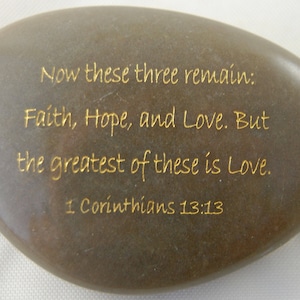 May include: A smooth, oval-shaped stone with gold lettering. The text reads: "Now these three remain: Faith, Hope, and Love. But the greatest of these is Love. 1 Corinthians 13:13."