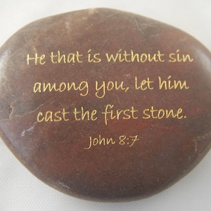 May include: A smooth, brown stone with gold lettering. The text reads, "He that is without sin among you, let him cast the first stone. John 8:7." The stone has a natural, organic shape.