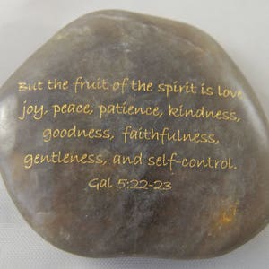 May include: A smooth, gray stone with gold lettering. The text reads: "But the fruit of the spirit is love, joy, peace, patience, kindness, goodness, faithfulness, gentleness, and self-control. Gal 5:22-23."