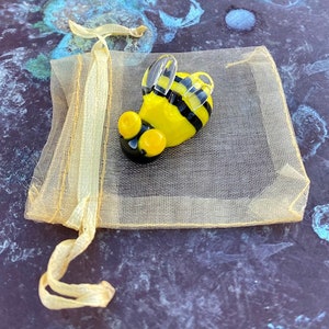 May include: A small glass bee figurine with yellow and black stripes, clear wings, and large black eyes. The bee is sitting in a yellow mesh bag.