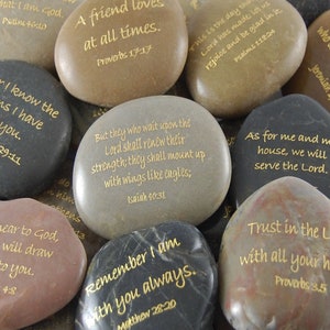 May include: A collection of smooth, natural stones with inspirational quotes and Bible verses in gold lettering. The stones vary in color from light brown to dark gray. The text includes phrases like "Trust in the Lord" and "A friend loves at all times."