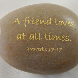 May include: A smooth, oval-shaped stone with the inscription "A friend loves at all times." in gold lettering. The text is followed by "Proverbs 17:17." The stone has a natural, earthy color.