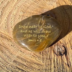 May include: A smooth, oval-shaped stone with the inscription "Draw near to God, and he will draw near to you. James 4:8" in gold lettering. The stone is a mix of brown and tan hues and rests on a textured wooden surface.