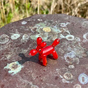 K&ouml;nnte beinhalten: Eine kleine, rote Glasfigur eines Hundes mit rundem K&ouml;rper, langen Ohren und einer knolligen Schnauze. Der Hund steht auf vier Beinen. Die Figur ist etwa 5 cm lang und vor einem unscharfen Hintergrund platziert.
