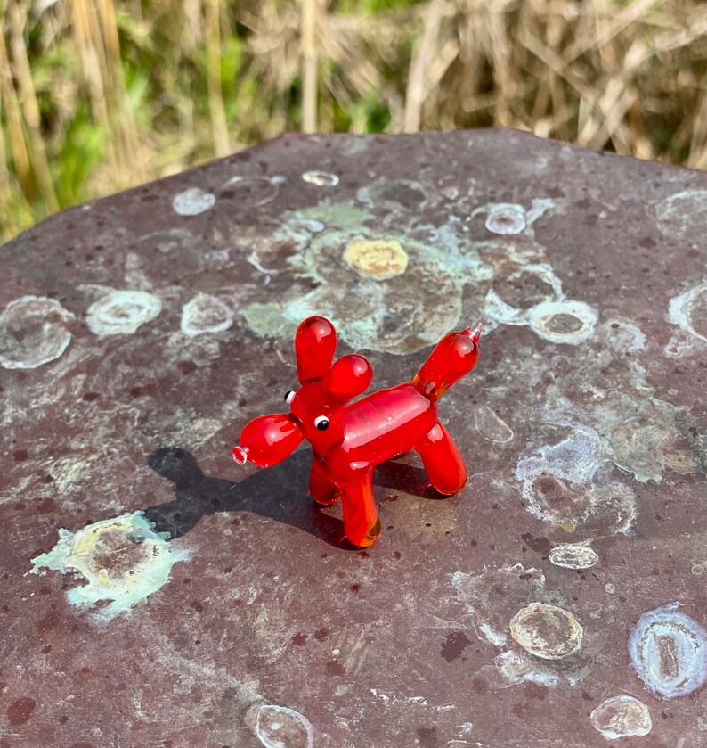 K&ouml;nnte beinhalten: Eine kleine, rote Glasfigur eines Hundes mit rundem K&ouml;rper, langen Ohren und einer knolligen Schnauze. Der Hund steht auf vier Beinen. Die Figur ist etwa 5 cm lang und vor einem unscharfen Hintergrund platziert.