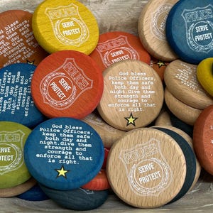 Op de afbeelding: Een verzameling houten fiches in verschillende kleuren, waaronder geel, oranje, blauw en natuurlijke houttinten. Elke fiche bevat de woorden "POLICE SERVE PROTECT" en sommige hebben een gebed. De fiches zijn rond en hebben een schildontwerp.