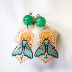 May include: A pair of earrings featuring a moth design. Each earring has a green bead at the top, with a silver hook. The moth design is teal and orange, set against a yellow sun and a cream-colored background.