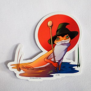 May include: A sticker featuring a cartoon illustration of a frog wearing a witch's hat and holding a staff. The frog is orange and green and is standing in a blue pond with green reeds. The background is a red circle.