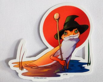 May include: A sticker featuring a cartoon illustration of a frog wearing a witch's hat and holding a staff. The frog is orange and green and is standing in a blue pond with green reeds. The background is a red circle.