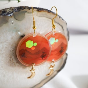 May include: A pair of gold hoop earrings with a red and orange sunset scene and geometric shapes. The earrings have a crescent moon charm hanging from the bottom.