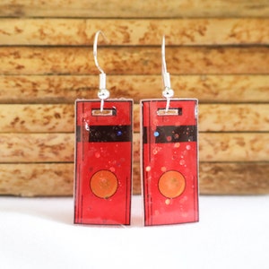 May include: A pair of rectangular earrings with a red background, black stripes, and an orange circle. The earrings have silver hooks and are decorated with glitter.