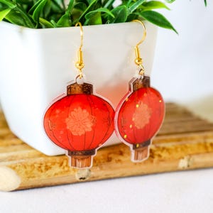 May include: Red and gold Chinese lantern earrings with gold hooks. The lanterns have a white floral design and are made of clear acrylic.