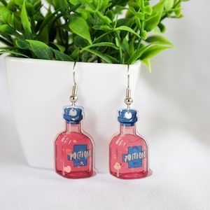 May include: A pair of silver dangle earrings featuring pink potion bottles with blue tops and the word "POTION" printed on the front. The bottles have a glittery effect.