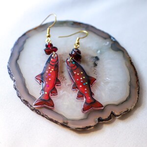 May include: A pair of fish-shaped earrings with red bodies and gold-colored hooks. The earrings have a glittery design with colorful dots and are adorned with small, dark red beads. The earrings are displayed on a white and gray stone.
