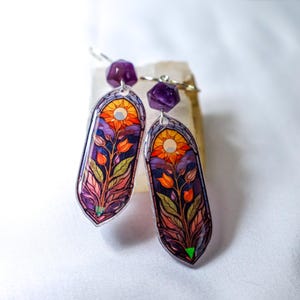May include: A pair of dangle earrings featuring elongated, arched pendants with a stained-glass style print of orange flowers, green leaves, and a yellow sun against a purple sky. Each earring is topped with a faceted, deep purple bead.
