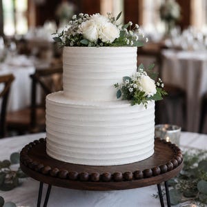 May include: A two-tier white cake with horizontal textured frosting, topped with white roses and baby's breath. A smaller cluster of white roses and greenery adorns the side. The cake sits on a dark brown wooden stand with a beaded edge and metal legs.