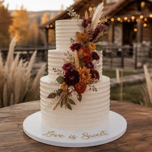May include: A three-tiered white cake with horizontal ridged frosting, decorated with a cascading arrangement of dried flowers in shades of burgundy, orange, and cream. The white cake base features the gold script text Love is Sweet.