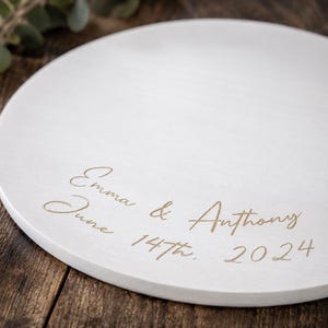 May include: A round, white, flat decorative plate featuring gold-colored engraved text that reads Emma & Anthony, June 14th, 2024. The surface is smooth and circular, with the personalized inscription centered in a cursive, elegant script font.