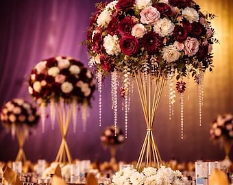 May include: Elegant floral centerpieces with burgundy, white, and pink roses and dahlias. The arrangements are set on tall, gold stands, with cascading crystal accents. The tables are set for a formal event, with gold napkins and glassware.