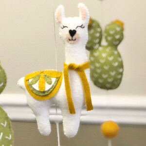 May include: A white felt llama with a yellow felt saddle and scarf hanging from a mobile. The llama has black eyes and a pink nose. The mobile also includes green felt cacti with white hearts.