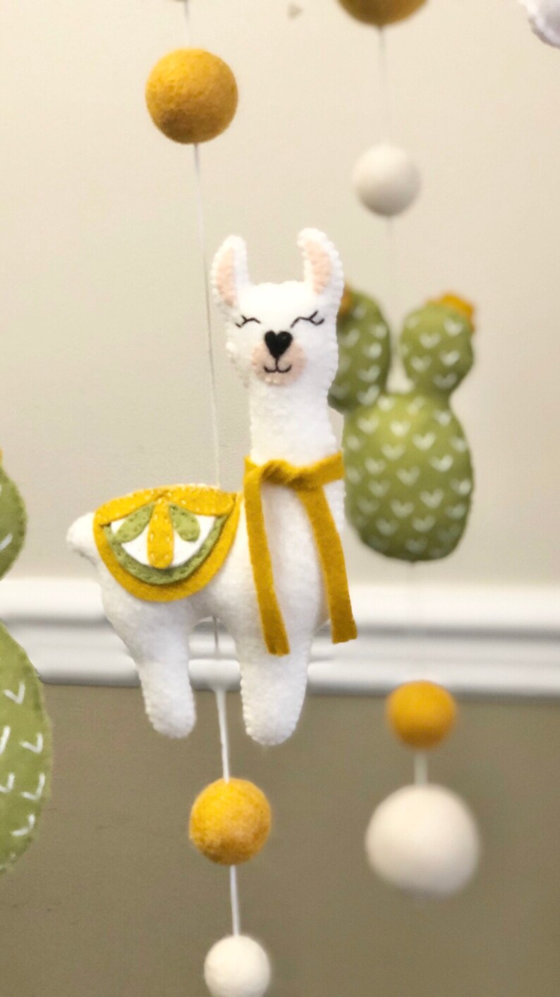 May include: A white felt llama with a yellow felt saddle and scarf hanging from a mobile. The llama has black eyes and a pink nose. The mobile also includes green felt cacti with white hearts.