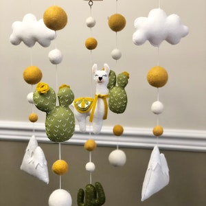 May include: A white, green, and yellow felt baby mobile with a llama, cactus, clouds, and mountains. The mobile is hanging from a wooden frame.