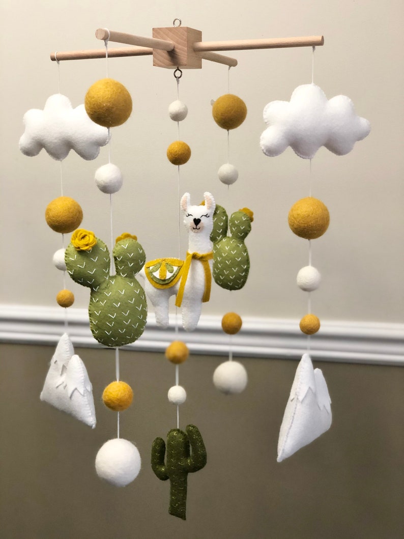 May include: A white, green, and yellow felt baby mobile with a llama, cactus, clouds, and mountains. The mobile is hanging from a wooden frame.