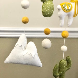May include: A felt baby mobile with a white mountain, two green cacti, a white llama, and yellow and white balls.
