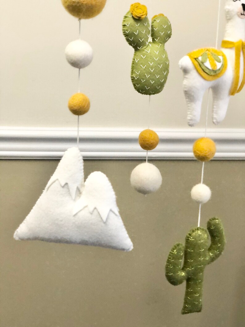 May include: A felt baby mobile with a white mountain, two green cacti, a white llama, and yellow and white balls.
