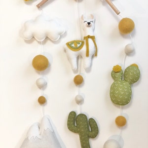 May include: A white felt llama with a yellow felt saddle and a yellow felt ribbon hangs from a wooden mobile. The mobile also features a white felt cloud, two green felt cacti, a white felt mountain, and yellow and white felt balls.
