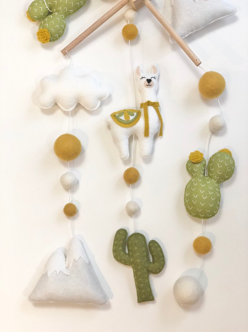 May include: A white felt llama with a yellow felt saddle and a yellow felt ribbon hangs from a wooden mobile. The mobile also features a white felt cloud, two green felt cacti, a white felt mountain, and yellow and white felt balls.