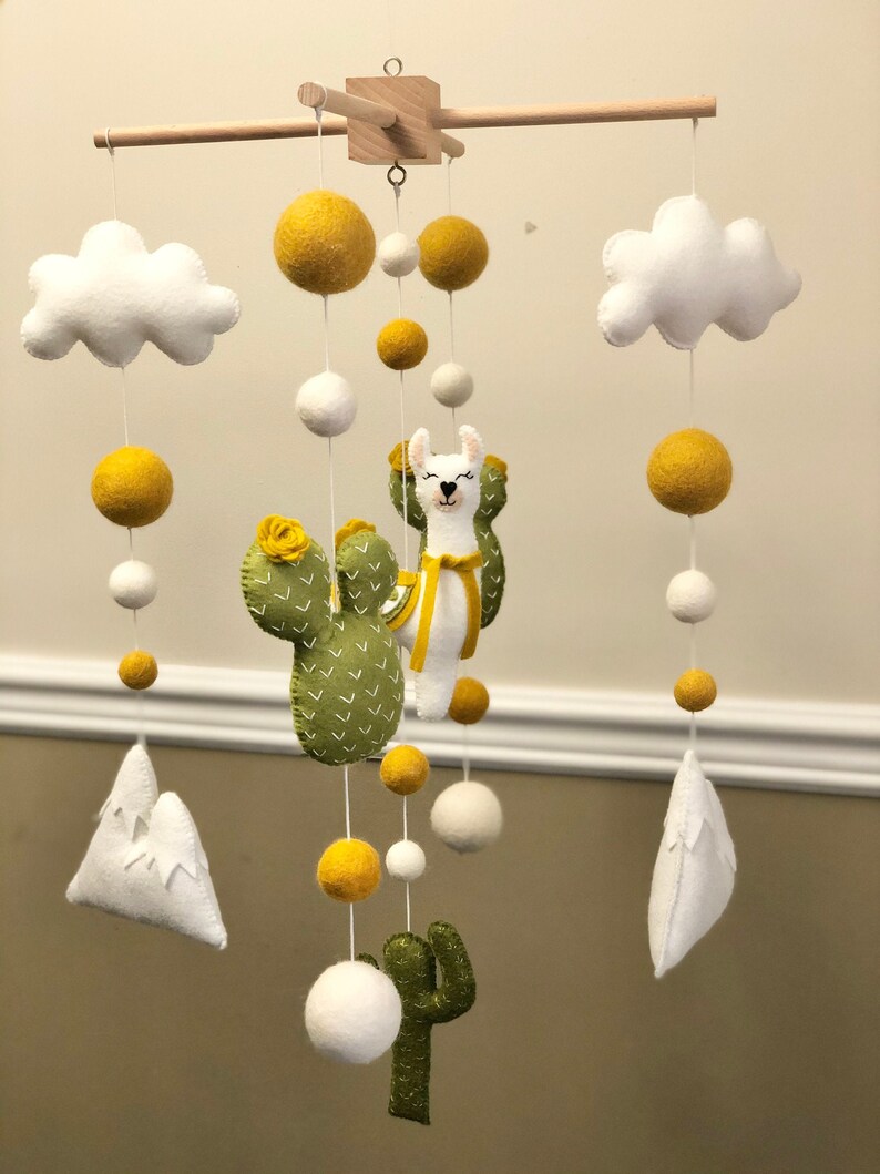 May include: A baby mobile with a llama, cactus, and clouds. The mobile is made of felt and has yellow and white pom-poms.