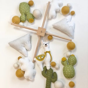 May include: A white, green, and yellow felt baby mobile with a llama, cacti, and clouds. The mobile is suspended from a wooden dowel.