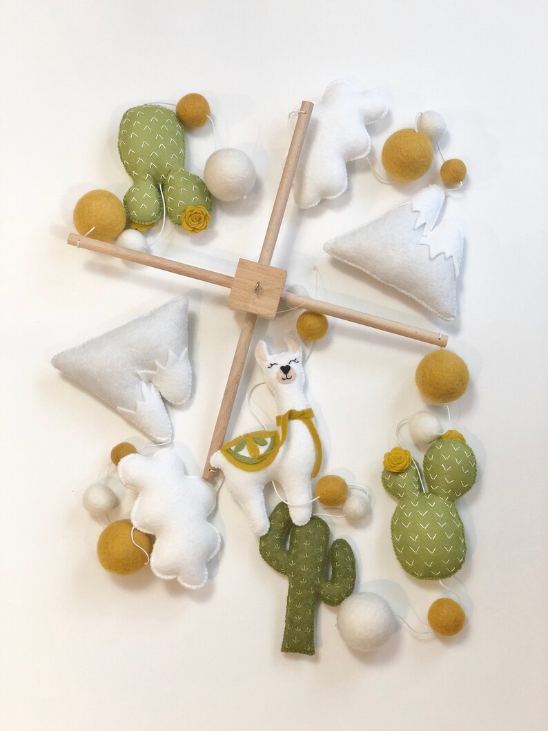 May include: A white, green, and yellow felt baby mobile with a llama, cacti, and clouds. The mobile is suspended from a wooden dowel.