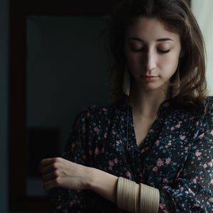 May include: A person wearing a black floral dress and wooden bangles. The dress has a V-neck and long sleeves. The bangles are light-coloured and stacked on the wrist. The background is a dark room with a mirror.