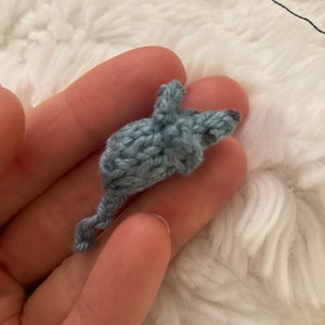 May include: A small, crocheted mouse in shades of blue. The handmade toy features a detailed design with a long tail and small ears. The mouse is held in a hand against a fluffy white background.