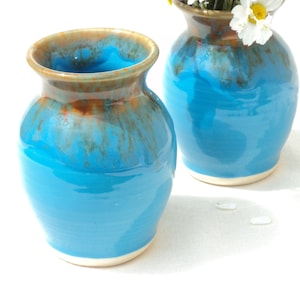 May include: Two blue ceramic vases with a white rim and a brown glaze. The vases are on a white surface and one vase has a bouquet of white daisies.
