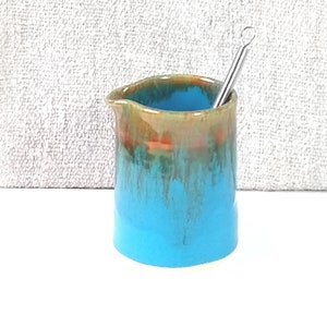 May include: A small, turquoise ceramic creamer with a brown and orange glaze. A metal stirring rod is placed inside the creamer.