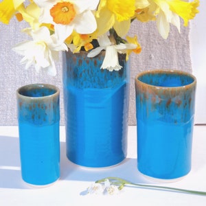 May include: A display of yellow and white daffodils in three turquoise ceramic vases. The vases have a glossy finish with a brown, dripped glaze at the rim. A few daffodils are placed near the vases.