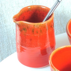 May include: A bright orange ceramic creamer with a metal spring inside. The creamer has a glossy finish and a slight green glaze.