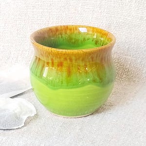 May include: A small, rounded ceramic cup featuring a vibrant lime green base that transitions into a warm, honey-brown glazed rim. The surface has a smooth, glossy finish with subtle vertical drip patterns where the two colours meet at the top.