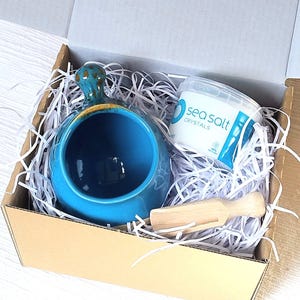 May include: A blue ceramic salt cellar with a wooden scoop and a container of sea salt crystals. The salt cellar has a textured glaze and a handle. The container of sea salt crystals has a white label with the text "sea salt crystals" and a blue circle with a white wave design.